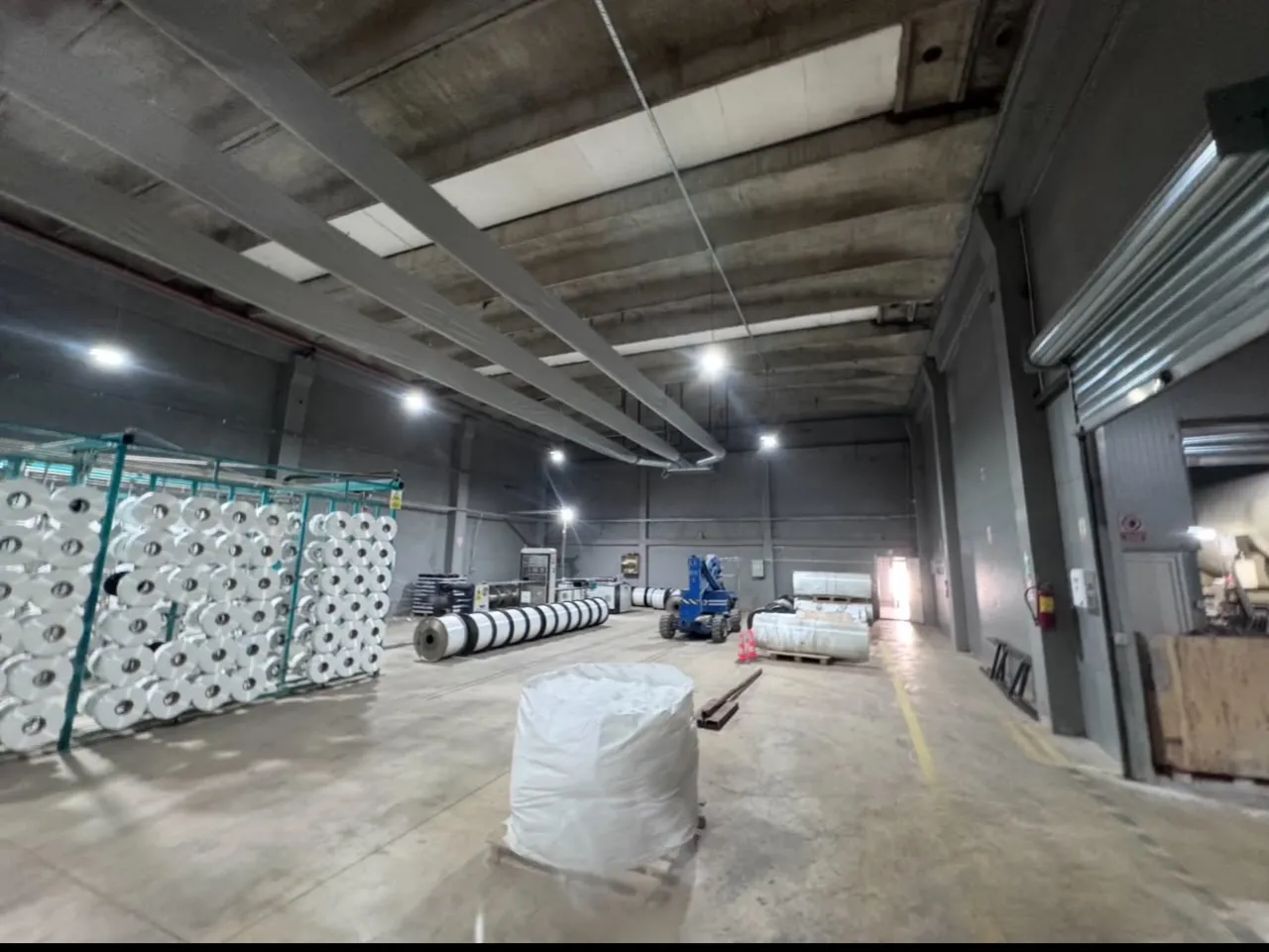 Hală industrială înaltă, cu iluminat pe grinzi, tubulatură la tavan și role mari în fundal
