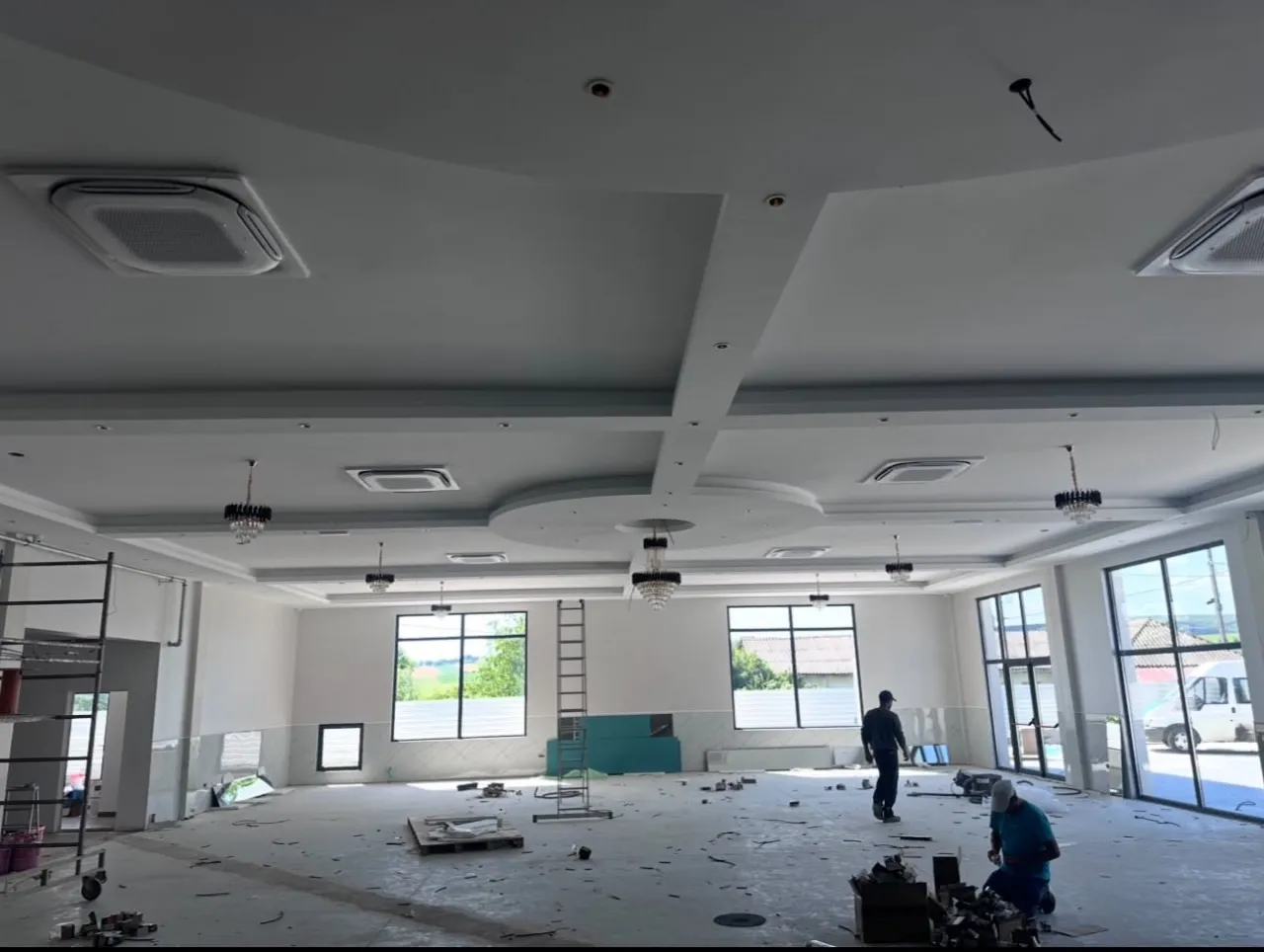 Sală mare în construcție, cu casete de climatizare în tavanul fals și doi oameni lucrând în fundal