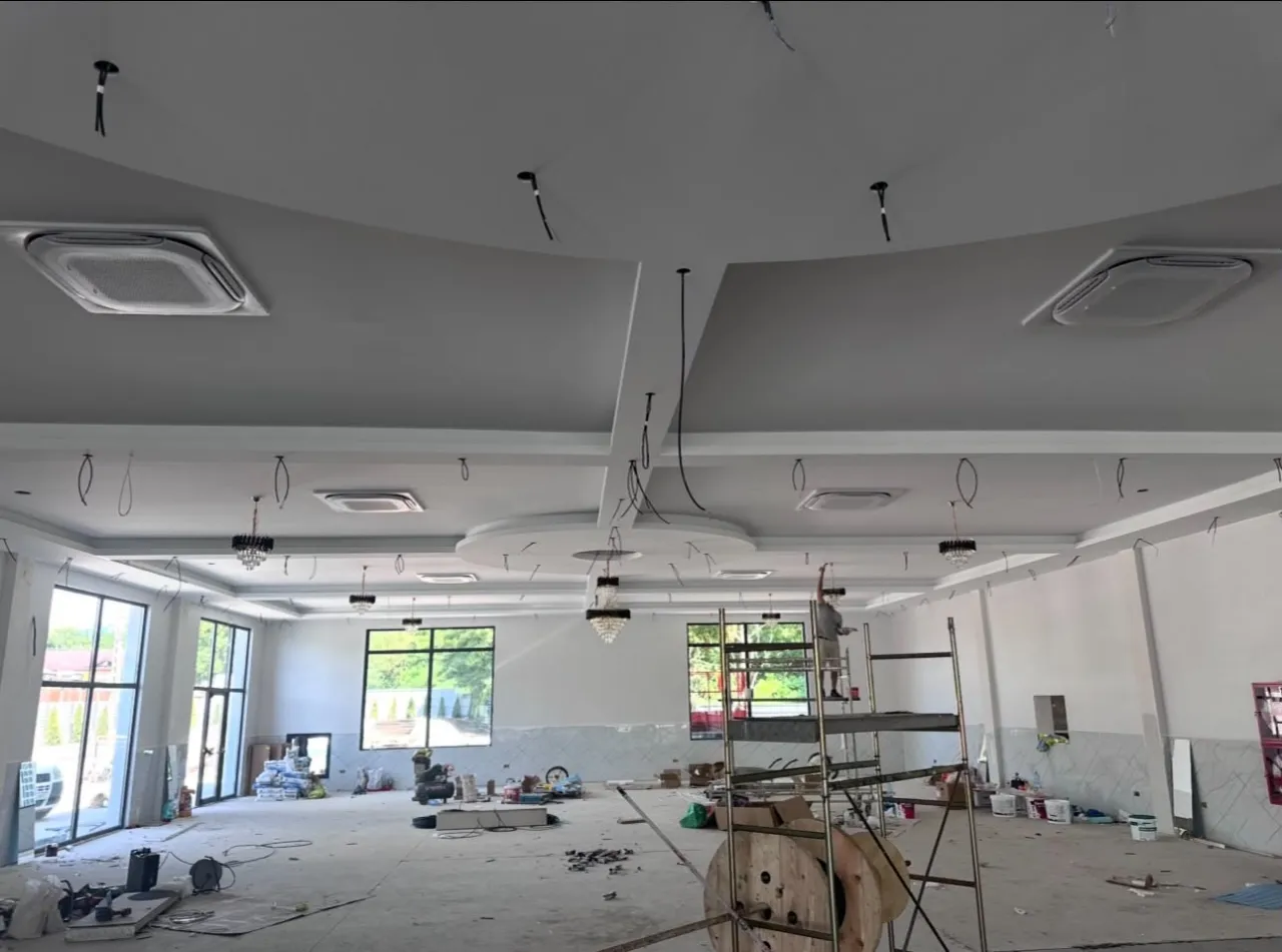 Aceeași sală în construcție, cu schelă lângă ferestre, casete de climatizare în tavan și o bobină de cablu pe podea
