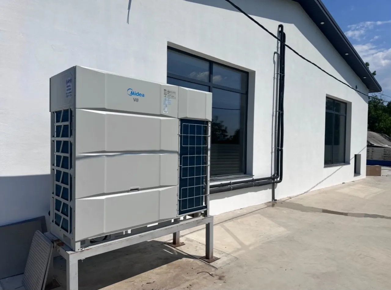 Unitate exterioară Midea VRF pe suport, lângă o clădire albă, cu țevi izolate negre pe fațadă